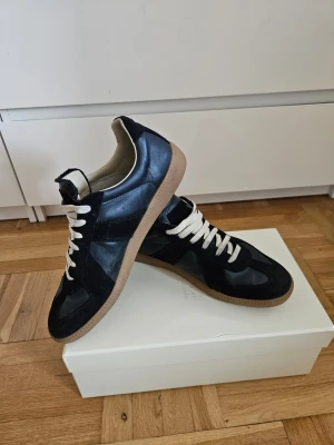 Maison Margiela Navy Gats - Stilrena Maison Margiela mörkblå sneakers med beige snören och brun sula. Skorna har en kombination av slätt läder och mocka på ovandelen, vilket ger en snygg kontrast. Låg modell som passar perfekt till jeans eller streetwear. Klassisk siluett och tidlös design.