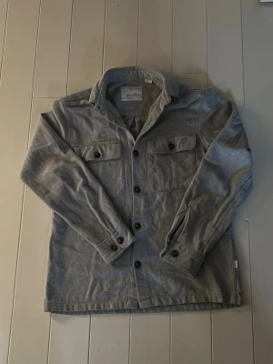 Grå overshirt från jack & jones - Snygg grå overshirt i mjukt tyg med två bröstfickor med lock och knappar. Klassisk krage och knäppning framtill. Snyggt lager på lager med t.ex en hoodie under. Perfekt till hösten/vintern om man inte vill ha en tjock jacka på sig när det är kallt ute.