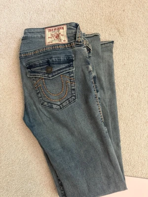 Blå bootcut jeans True Religion - Säljer ett par blå bootcut jeans från True Religion med slitningar och trasiga detaljer på ena benet. Jeansen har klassiska kontrastsömmar och ficklock bak med knapp. Perfekta för dig som gillar en avslappnad och trendig stil. Storleken är 27 motsvarar typ s/m🥰Jeansen har en missfärgad fläck på bakre benet, därav sjunker värdet 