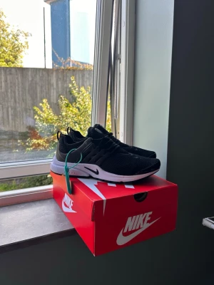 Svarta Nike sneakers med vit sula - Snygga svarta Nike sneakers med vit sula och svart mesh-överdel. Skorna har en låg profil och klassisk Nike-logga på sidan. Perfekta för dig som gillar stilrena och sportiga sneakers med bekväm passform.