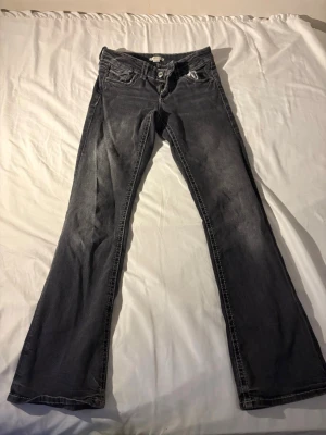 Grå bootcut jeans med fickdetaljer - Snygga grå bootcut jeans med coola fickdetaljer bak. Jeansen har klassisk femficksmodell framtill och två bakfickor med lock och knapp. Perfekta för en avslappnad och trendig look.