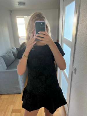 Svart kort klänning med volang - Säljer en svart kort klänning med volangkant nedtill och diskret blommönster. Klänningen har korta ärmar och en rund halsringning. Materialet ser ut att vara polyester eller liknande och ger en elegant känsla. Oanvänd!!!!!