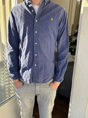Blå Ralph lauren skjorta - Säljer en klassisk blå ralph Lauren skjorta med knappar framtill och button-down krage. Skjortan har lång ärm och en gul broderad logga på bröstet. Tillverkad i mjuk bomull och passar perfekt till jeans eller chinos. Pris går att diskutera!!