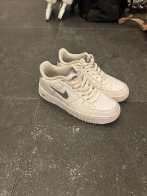 Nike Air Force 1 vita sneakers - Klassiska vita Nike Air Force 1 sneakers med lågt skaft och silverfärgad swoosh. Skorna har perforerad tå för ventilation, vita snören och en robust sula. Tillverkade i läder med textildetaljer och ikonisk Air-dämpning i sulan.