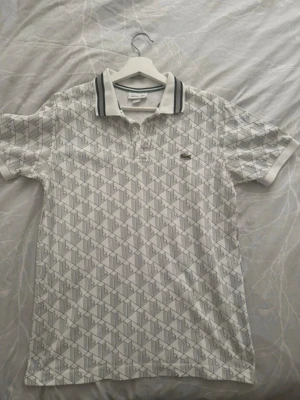 Vit mönstrad piké från Lacoste - Säljer en vit pikétröja från Lacoste med grått geometriskt mönster och klassisk krage med gröna och svarta ränder. Tröjan har korta ärmar och den ikoniska krokodilloggan på bröstet. Perfekt för en clean och sportig look.