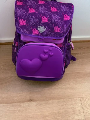 Lila ryggsäck med hjärtmotiv - Säljer en lila ryggsäck från med rosa och lila mönster av klossar och hjärtan. Framtill finns ett stort hjärtmotiv i relief. Väskan har flera fack, dragkedjor och plats för namn och telefonnummer på insidan. Perfekt för skolan eller utflykter.