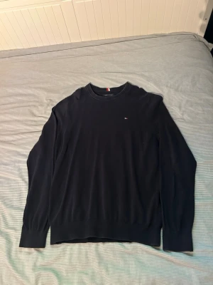 Blå tröja från Tommy Hilfiger - Klassisk marinblå tröja från Tommy Hilfiger med rund halsringning och diskret logga på bröstet. Tröjan har ribbade muddar vid ärmslut och nederkant. Perfekt för en clean och stilren look.