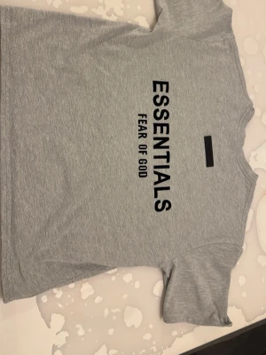 Grå Essentials Fear of God t-shirt - Snygg grå t-shirt från Essentials Fear of God med svart tryck både fram och bak. Klassisk passform och rund hals. Perfekt till streetwear-stilen och enkel att matcha med allt. Tillverkad i mjuk bomull för skön känsla hela dagen.
