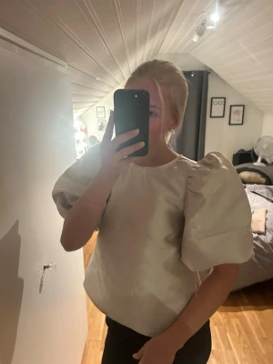 Vit topp med puffärmar - Säljer en stilren vit topp med stora puffärmar och rund hals. Toppen har en lätt glansig finish och är perfekt för dig som vill sticka ut med en trendig siluett. Använd en gång så är nästintill oanvänd. Strl xs