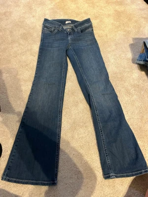 Blå bootcut jeans  - Säljer ett par klassiska blå lågmidjade bootcut jeans med låg midja och snygga fickdetaljer bak. Jeansen är stretchiga och har några defekter längst ner på byxbenen och ett hål i fickan (se bilder)