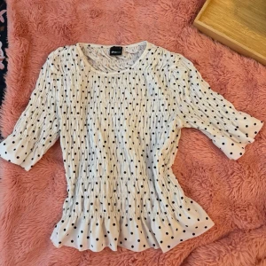 Vit prickig blus med volang - Supersöt vit blus med svarta prickar och volangkant nertill. Blusen har trekvartsärm och smockdetaljer som ger en snygg passform. Perfekt att styla med jeans eller kjol för en trendig look.