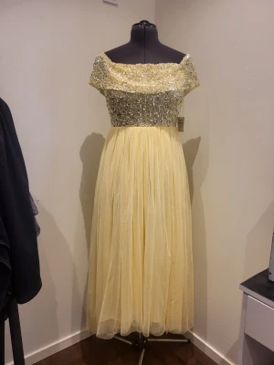 Gul axelbandslös långklänning - Elegant gul långklänning med axelbandslös design och glittrande paljetter över bysten. Kjolen är i flera lager tunn tyll som ger ett svepande intryck. Perfekt för bal eller festliga tillfällen där du vill sticka ut. Stl 38, bröst ca 97-100cm, längd ca 112 från armhåla. Strechigt tyg. Aldrig använd.
