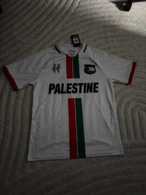 Vit Palestina fotbollströja - Säljer en vit fotbollströja med texten 'PALESTINE' på bröstet och ränder i rött och grönt. Tröjan har röda muddar och detaljer samt emblem och karta på både fram- och baksidan. Perfekt för dig som vill visa ditt stöd på planen eller läktaren.
