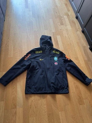 Svart Brasilien Nike vindjacka - Svart vindjacka med huva från Nike, prydd med Brasilien fotbollslandslagets logga och flera sponsorloggor i gult, vitt och rött. Jackan har dragkedja framtill, fickor och justerbar huva. Perfekt för dig som gillar sportig stil och vill sticka ut. den är xl men passar för m och l.Mer bilder kommer snart för jag är på semester nu .