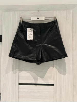 ZARA Faux Leather Shorts - Helt nya och superstylisiga svarta shorts i konstläder från ZARA. Perfekt passform med hög midja, fickor och en snygg finish. Passar lika bra till boots och stickat som till klackar på festen!  Märke: ZARA  Storlek: S (USA S / MEX 26)  Skick: Helt nya med prislapp kvar (NWT)  Material: Faux leather (konstläder) med mjuk insida  Mått: Midja ca 35–36 cm, längd ca 35 cm 🚫 Hem: Rök- och djurfritt hem 🚚 Snabb leverans: Skickar inom 24 timmar! 