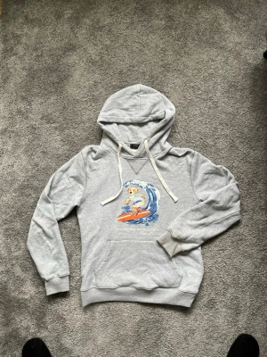 Grå hoodie med surfande björn - Säljer en ljusgrå hoodie med en cool print av en björn som surfar på framsidan. Hoodien har snörning i huvan och en klassisk känguruficka. Perfekt för dig som gillar chill och streetwear vibes.