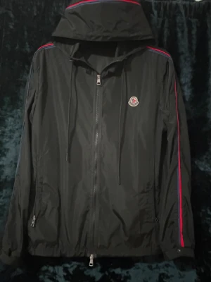 Moncler vind jacka  - Snygg svart vindjacka med huva och dragkedja framtill. Jackan har röda och blå ränder längs ärmarna. Tillverkad i lätt, vindtätt material och har fickor med dragkedja.