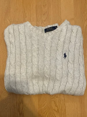 Polo Ralpn Lauren Knitted Sweater  - Säljer en klassisk Polo Ralph Lauren Cable Knit i snygg grå färg med den ikoniska mörkblå Polo loggan på bröstet.  Väldigt stilren och lätt att matcha – perfekt för en klassisk old money/preppy look, men fungerar lika bra till vardags.  • Polo Ralph Lauren • Storlek M • Klassisk Cable Knit • Bra skick  Hör gärna av dig vid frågor eller bud!  