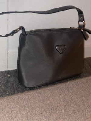 Svart axelremsväska i nylon - Snygg svart handväska i nylon med axelrem och dragkedja upptill. Väskan har en stilren, rektangulär form och ett litet trekantigt emblem framtill. Perfekt storlek för det viktigaste och enkel att matcha med olika outfits.