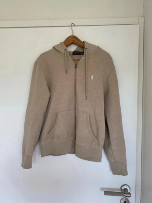 Polo Ralph Lauren hoodie - Snygg beige hoodie från Polo Ralph Lauren med dragkedja framtill, två stora fickor och klassisk huva med dragsko. Liten vit logga broderad på bröstet. Tillverkad i mjukt bomullsmaterial som känns skönt mot huden. Perfekt för en avslappnad och stilren look. Säljer för att den inte andvänds längre