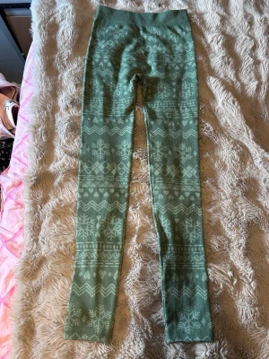 Gröna leggings med vintermönster - Snygga gröna leggings med stickat vintermönster i ljusare grönt. Mönstret består av snöflingor och geometriska former längs benen. Hög midja och stretchigt material som sitter skönt. Perfekta för kalla dagar och mysiga hemmakvällar.