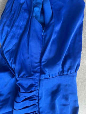 Lång blå klänning med drapering - Snygg långklänning i klarblått med draperad detalj på sidan och rund halsringning. Klänningen är ärmlös och har en slät, elegant yta som ger ett lyxigt intryck. Perfekt för dig som vill sticka ut med färg och stil.