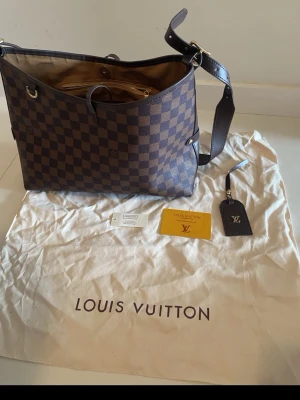 Louis Vuitton Damier med kvitto  - Säljer en brun handväska från Louis Vuitton i klassiskt rutigt Damier-mönster. Väskan har justerbar axelrem, guldfärgade metalldetaljer och beige insida. Kommer med dustbag, tag och kvitto. 