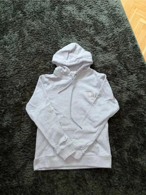 Ljusgrå hoodie från D10R - Säljer en ljusgrå hoodie med huva och dragsnören. Tröjan har en broderad logga på bröstet och en klassisk känguruficka framtill. Tillverkad i mjukt sweatshirtmaterial som är perfekt för chill dagar. Långa ärmar med ribbade muddar och avslappnad passform. Pris kan diskuteras vid SBAB affär 