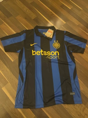 Inter Milan matchtröja blå/svart Nike - Snygg Inter Milan matchtröja från Nike i blått och svart med gula detaljer. Tröjan har klassiska vertikala ränder, korta ärmar och krage. På bröstet finns klubbmärke, Nike-logga och sponsortryck i gult. Perfekt för fotbollsfans!