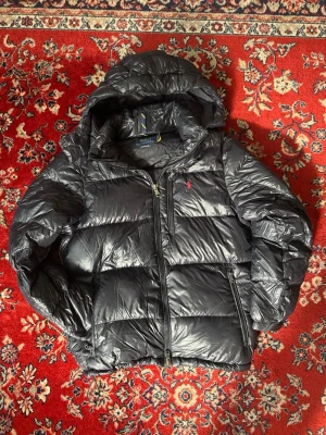 Svart dunjacka från Polo Ralph Lauren - Snygg svart dunjacka från Polo Ralph Lauren med huva och dragkedja framtill. Jackan har två sidofickor med dragkedja och den klassiska röda loggan broderad på bröstet. Perfekt för kalla dagar och riktigt skön att bära.