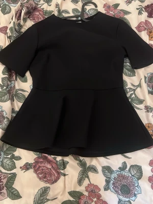 Svart peplum topp med kort ärm - Snygg svart peplum topp med rund halsringning och korta ärmar. Toppen har en markerad midja och utställd nederdel som ger en feminin siluett. Perfekt att styla med jeans eller kjol för en trendig look.