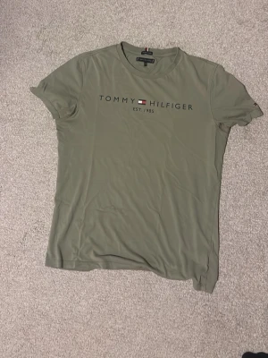 Olivgrön t-shirt från Tommy Hilfiger - Snygg olivgrön t-shirt från Tommy Hilfiger med tryckt logga och text på bröstet. Klassisk rund halsringning och korta ärmar. Perfekt till jeans eller shorts för en clean och enkel look.