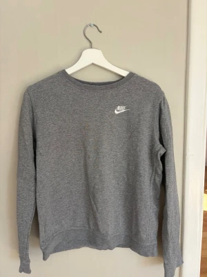 Grå sweatshirt från Nike - Klassisk grå sweatshirt från Nike med rund halsringning och vit broderad logga på bröstet. Tröjan har långa ärmar och ribbade muddar vid ärmslut och nederkant. Perfekt för en avslappnad och sportig stil.