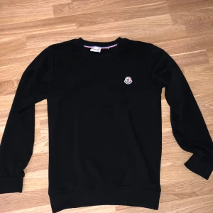 Svart tröja från Moncler - Säljer en stilren svart tröja från Moncler i storlek M. Tröjan har en broderad Moncler-logga på bröstet och är tillverkad i ett mjukt material. Perfekt för både vardag och fest!