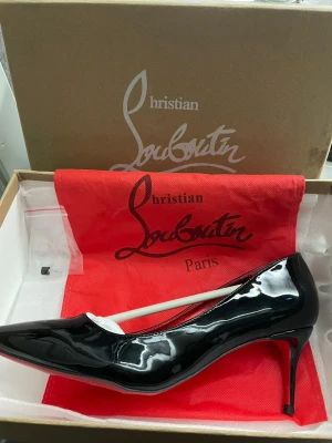 Svarta lackpumps från Christian Louboutin - Säljer ett par klassiska svarta pumps i lack från Christian Louboutin. Skorna har spetsig tå, röd innersula och den ikoniska röda sulan. Klacken är smal och elegant, och materialet är glansigt lackläder. Perfekta för dig som vill sticka ut med stil. Lätt använda annars unga problem med de har lådan och alla tillbehör