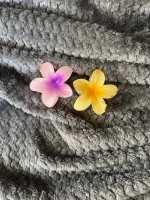 Blommiga hårklämmor i plast - Två hårklämmor i form av blommor, en i rosa och lila och en i gul och orange. Båda är tillverkade i glansig plast och har en tydlig blomform med fem kronblad. Perfekta för att ge en färgglad touch till din frisyr.