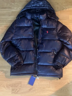 Mörkblå dunjacka från Polo Ralph Lauren - Snygg mörkblå dunjacka från Polo Ralph Lauren med huva och dragkedja. Jackan har en röd logga på bröstet, stora vadderade partier och praktiska fickor med dragkedja. Perfekt för kalla vinterdagar och riktigt trendig look.