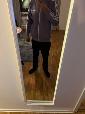 Lila zip-tröja från Palm Angels - Säljer en lila zip-tröja från Palm Angels med vita ränder längs ärmarna och vit dragkedja framtill. Tröjan har en sportig look med ribbade muddar och hög krage. Perfekt för dig som gillar streetwear och vill sticka ut. Knappt använd 