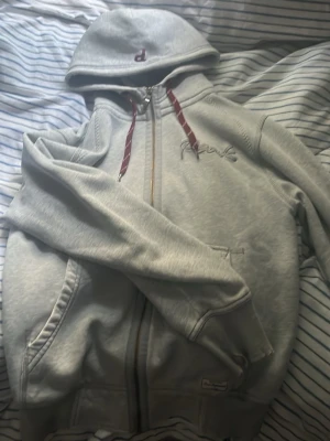 Ljusgrå hoodie med dragkedja från peakpreformance - Ljusgrå hoodie med dragkedja och fickor framtill. Har huva med röda snören och ett broderat 'P' på huvan samt broderad text på bröstet. Tillverkad i mjukt sweatshirtmaterial, perfekt för chill dagar.