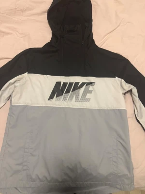Nike svart och grå vindjacka - Nike vindjacka i svart och ljusgrått med stor logga framtill. Jackan har huva med dragkedja, raka ärmar och två sidofickor. Tillverkad i lätt, vindtätt material som passar perfekt för blåsiga dagar.