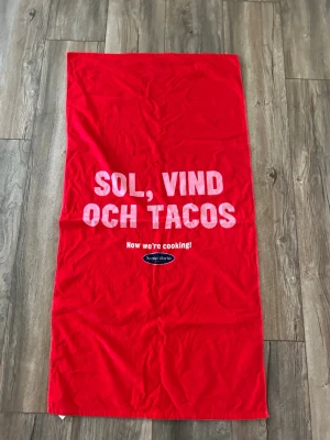 Röd strandhandduk Sol, Vind och Tacos - Röd strandhandduk med texten 'Sol, vind och tacos' i vitt och rosa samt Santa Maria-logga. Handduken är rektangulär och har en mjuk frottéliknande baksida i vitt. Perfekt för stranden eller poolen.
