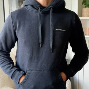 Svart hoodie från Peak Performance - Snygg svart hoodie från Peak Performance med klassisk huva och dragsnören. Har en stor magficka framtill och diskret logga på bröstet. Tillverkad i mjukt bomullsmaterial och har ribbade muddar vid ärmslut och nederkant. Perfekt för en avslappnad stil.