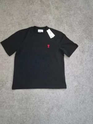 Svart t-shirt med röd detalj - En stilren svart t-shirt med korta ärmar och en liten röd broderad detalj på bröstet. T-shirten har en klassisk passform och rund halsringning. Perfekt basplagg för en enkel och clean look. Har tyvärr inte kvar lappen i bilderna. Är strl S men kan passa vissa som har M