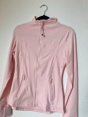 Lululemon pink  - Snygg ljusrosa sportjacka med hel dragkedja framtill och två fickor med dragkedja. Jackan har hög krage och är tillverkad i ett stretchigt, mjukt material som passar perfekt till träning eller promenader. Snygga sömdetaljer ger en sportig look.
