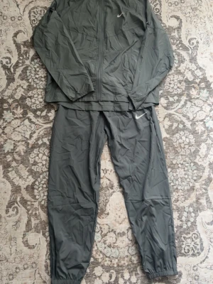 Nike Miller tracksuit - Säljer ett grått träningsset från Nike med jacka och byxor. Setet har dragkedja framtill på jackan, elastiska muddar på byxorna och diskreta vita Nike-loggor. Perfekt för löpning eller annan träning. Materialet är lätt och andas bra.