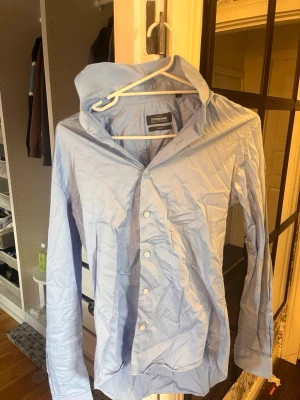 Ljusblå slim fit skjorta Dressmann - Snygg ljusblå skjorta från Dressmann i slim fit-modell. Tillverkad i stretchig bomull för extra komfort. Klassisk krage och knäppning framtill med vita knappar. Perfekt för en clean och stilren look. Skriv om ni har några frågor!