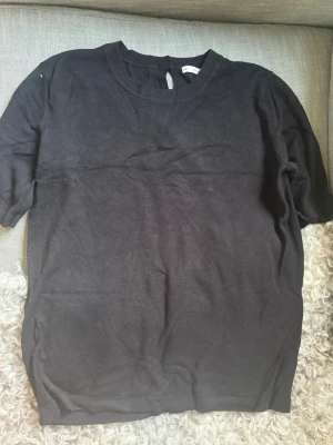Svart kortärmad topp - En enkel svart topp med rund halsringning och korta ärmar. Toppen har en knappdetalj i nacken och är tillverkad i ett mjukt, tunt material som känns bekvämt mot huden. Storlek är L men passar bra mig som brukar bära S