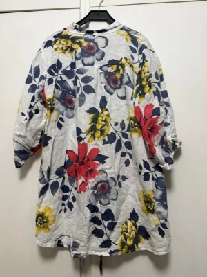 Vit blommig blus med knappar - Snygg vit blus med stora blommor i rött, gult och blått. Blusen har trekvartsärm, bröstficka och knäppning framtill. Tillverkad i ett lätt och luftigt tyg, perfekt för en avslappnad och trendig look.