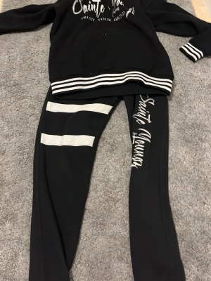 Svart set med vita detaljer - Säljer ett svart tvådelat set med sweatshirt och leggings. Setet har vita ränder på ärmslut och nederkant samt två vita ränder på ena benet. Texten 'Sainte Youngz' finns tryckt på både tröjan och byxan. Perfekt för en avslappnad och sportig look.