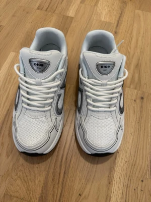 B30  - Säljer ett par vita sneakers med grå och svarta detaljer. Skorna har meshpaneler för extra ventilation,Snygg och modern design som sticker ut. Perfekta för dig som gillar exklusiva sneakers och streetwear.